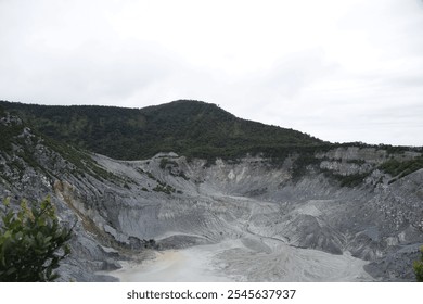 A panoramic view of a volcanic crater with a steep, rocky slope. The crater is filled with ash and surrounded by lush green vegetation. The sky is overcast, adding a dramatic atmosphere to the scene. - Powered by Shutterstock