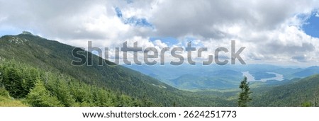Panoramic view from Whiteface Mountain in the Adirondacks, New York, featuring dramatic clouds, rolling green peaks, and the winding silhouette of Lake Placid in the valley below.