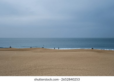 Panoramic view of a wide empty beach with gentle ocean waves and a few distant people standing near the shoreline under an overcast sky.