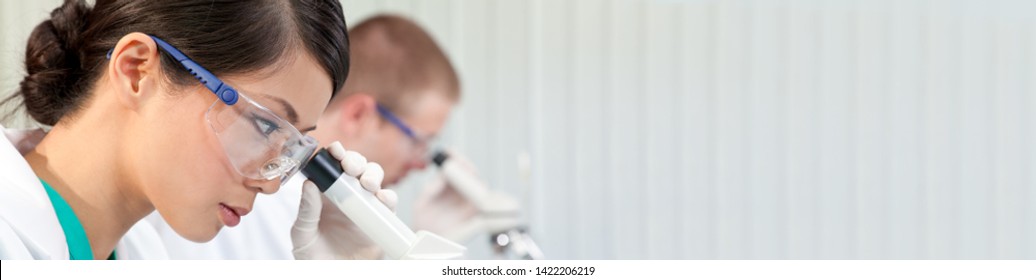 Panoramic Web Banner Of Chinese Asian Female Woman Scientist Researcher Or Doctor Using A Microscope In A Medical Research Lab Or Laboratory With Her Male Colleague Out Of Focus Behind Her.