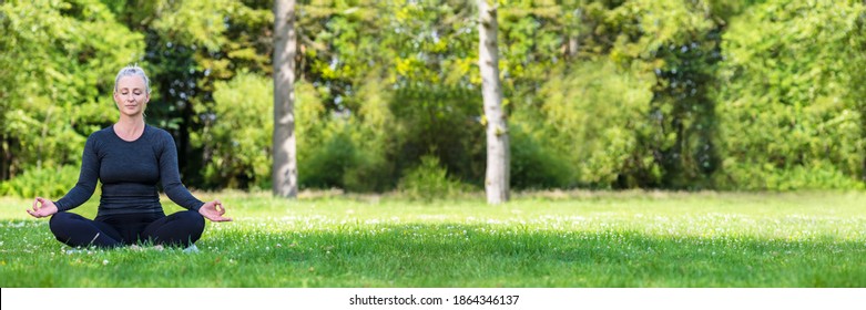 Panoramic web banner of mature middle aged fit healthy woman practicing yoga outsidein a natural tranquil green environment panorama.