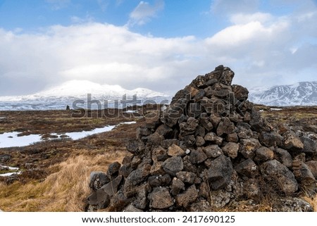 Panoramic winter landscape in Thingvellir National Park, Iceland with large stone cairn in foreground and white clouds over snowy and icy lava rock landscape and snowcapped mountains in background