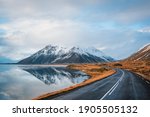 Panoramic winter photo of road leading along coast of lake to volcanic mountains. High rocky peaks covered with snow layer mirroring on water surface. Driver
