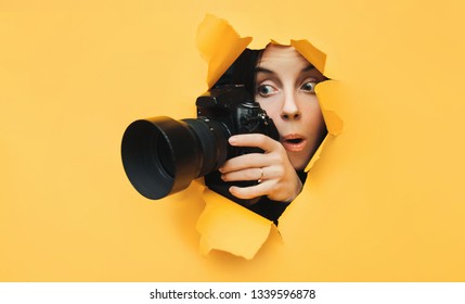 Ein paparazzi lustiges Mädchen mit einer Kamera schaut von vorne und sieht, was mit offenem Mund passiert. Gelbes Papier, zerrissenes Loch. Tablettenpresse. Auf der Suche nach dem Grundstück für Fotobestände.