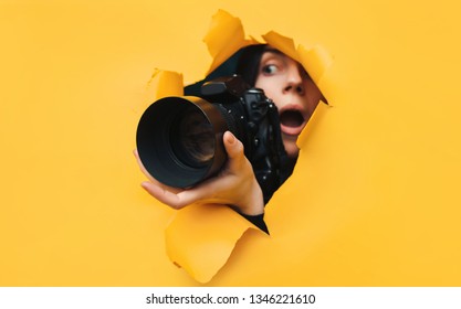 Ein Paparazzi-Mädchen mit einer schwarzen Reflexkamera schaut von vorne und sieht, was mit offenem Mund passiert. Gelbes Papier, zerrissenes Loch. Tablettenpresse. Auf der Suche nach dem Grundstück für Fotobestände.