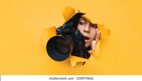 Ein paparazzi Mädchen mit einer Reflexkamera schaut von vorne und sieht, was mit offenem Mund passiert. Gelbes Papier, zerrissenes Loch. Tablettenpresse. Auf der Suche nach dem Grundstück für Fotobestände.