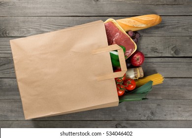 Paper bag full of different food on wooden background, close up. Grocery shopping concept, top view