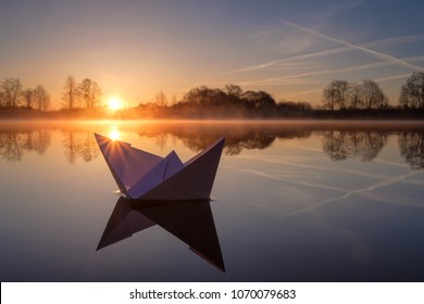 A Paper Boat Sailing Along The River At Dawn.