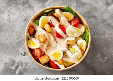 Paper Bowl With Caesar Salad Of Romaine Lettuce, Cherry Tomatoes, Quail Eggs, Croutons, Chicken And Sauce On Gray Surface