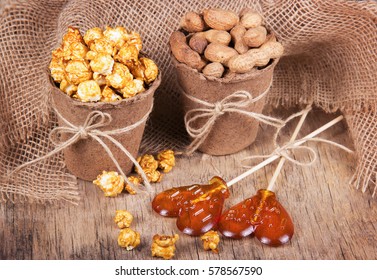 Paper Bucket Of Caramel Popcorn, Roasted Peanuts In Shell, And Lollipops. Set Meal For A Movie. Not Healthy Food And Snacks