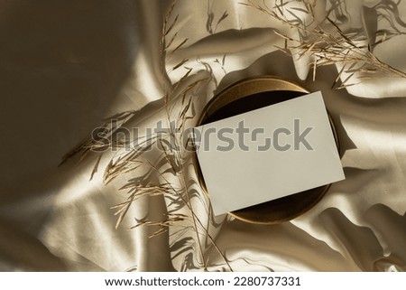 Paper card sheet with blank mockup copy space and dried grass on glossy gold silk cloth background. Aesthetic sunlight shadow silhouette. Luxury bohemian minimal invitation or business card template