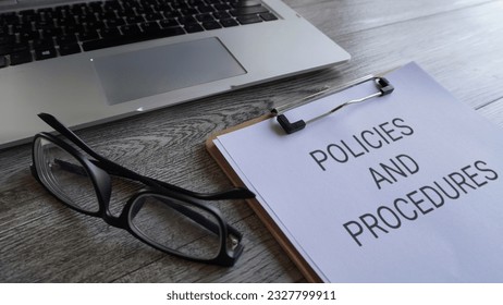 Paper clipboard with text POLICIES AND PROCEDURES on table with laptop and glasses.