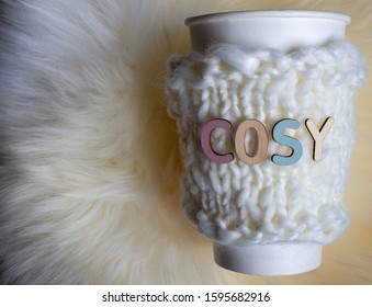 Paper Coffee Cup With Reusable Sleeve And Words Cosy On A Sheepskin Rug Background