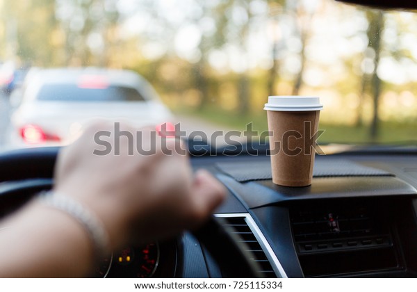 Paper Cup with coffee on the dashboard of the car\
blurred green background. Without focus the man\'s hand holds the\
steering wheel