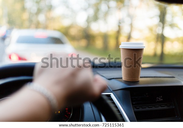 Paper Cup with coffee on the dashboard of the car
blurred green background. Without focus the man's hand holds the
steering wheel