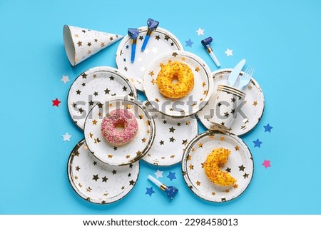 Paper disposable tableware with donuts, party blowers and hat on blue background
