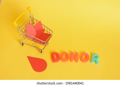 Paper Drops Of Blood And Shopping Cart And Inscription Donor On Yellow Background, World Donor Day Concept, Donate Blood For Money, Donor Blood Bank