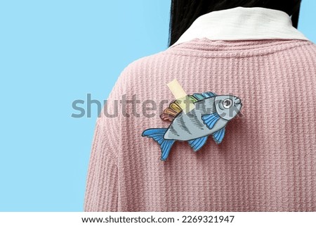 Paper fish on woman's back against blue background, closeup. April Fools' Day celebration