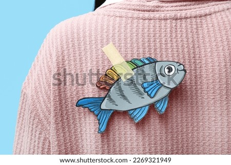 Paper fish on woman's back against blue background, closeup. April Fools' Day celebration