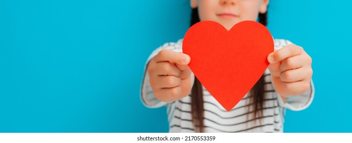 Paper Heart In Children's Hands. A Girl Child Hid Behind A Valentine. The Concept Of Recognition Of Love, Gratitude, Care, Health, Family.