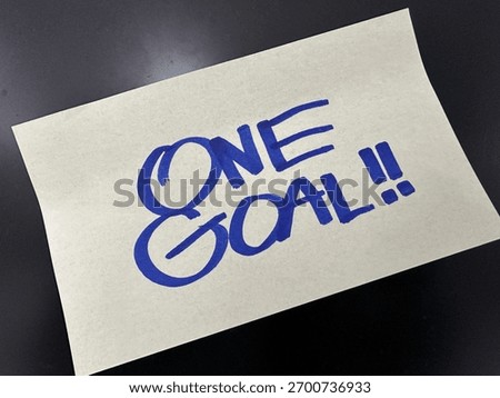 Paper note with bold blue text “ONE GOAL!!”—a motivational message emphasizing singular focus, determination, and purpose-driven action.

