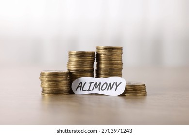 Paper note with word Alimony and stack of coins on wooden table, closeup