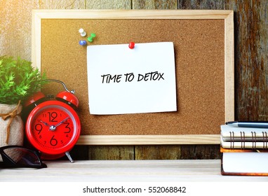 Paper note written with TIME TO DETOX inscription on cork board. Eye glasses, alarm clock, plant and books on wooden desk.