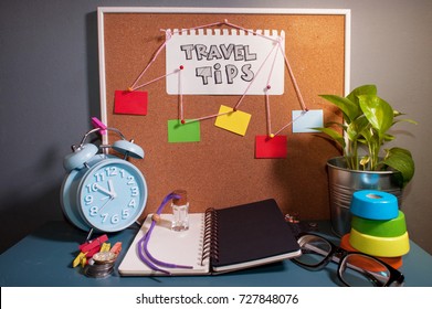 Paper Note Written With TRAVEL TIPS Inscription On Cork Board. Eye Glasses, Alarm Clock, Plant And Books On Wooden Desk. Artificial Grain Effect.