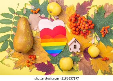 Paper Rainbow Heart LGBT Symbol And Wooden House Symbol Of Family On Multi-colored Red, Orange, Green Dry Fallen Autumn Leaves And Orange Rowan Berries, Apples And Pears On A Yellow Background.