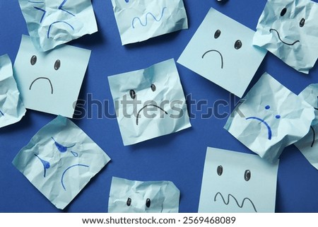 Paper sheets with drawn sad faces on blue background, top view