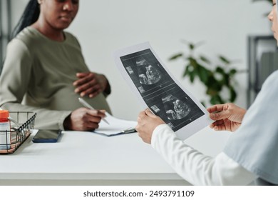 Paper with sonogram of fetus in hands of young gynecologist sitting by workplace in front of pregnant patient reading medical form - Powered by Shutterstock