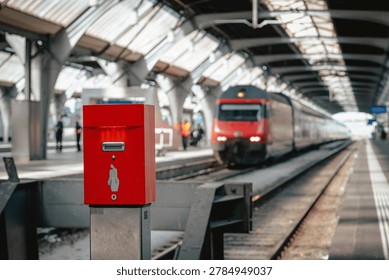Paper ticket validation machine located inside the Zurich Main Station (Zürich HB) in Switzerland, with local trains approaching at platform's entrance, modern Swiss railway public transportation
