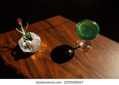 paper tulip in vase with green absinthe drink, cocktail on wooden table in the sun with harsh shadows