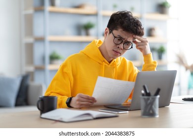 Paperwork Concept. Portrait Of Focused Asian Man In Eyeglasses Holding Reading Paper Or Financial Document, Sitting At Desk With Laptop Computer In Living Room, Thinking And Working At Home Office