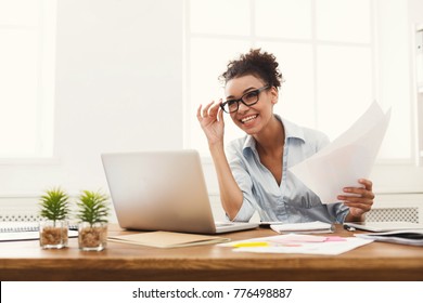 Paperwork. Happy Smiling African-american Business Woman In Formal Wear Sitting At Wooden Desk In Modern Office And Reading Report Document.