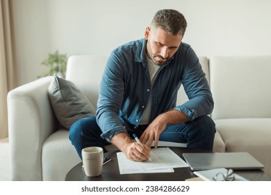 Paperwork. Middle aged man signing documents papers sitting on couch in living room indoors. Guy reviewing household documentation and writing business report at home - Powered by Shutterstock