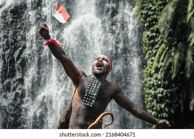 Papua man of Dani tribe say merdeka, celebrating Indonesia independence day against waterfall background.  - Shutterstock ID 2168404671