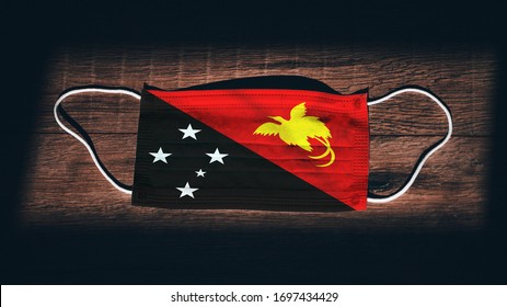 Papua New Guinea National Flag at medical, surgical, protection mask on black wooden background. Coronavirus Covid–19, Prevent infection, illness or flu. State of Emergency, Lockdown