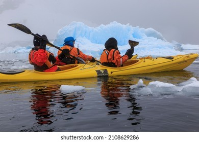 Paradise Bay, Antarctica - 12 09 17: Expedition Tour Guide In A Kayak On An Antarctic Kayaking Excursion  Guiding A Couple Of Cruise Kayak Tourists Next To A Blue Iceberg Floating In The Antarctic Sea