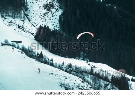 A paraglider soars over a snowy landscape, embodying the concept of freedom and adventure. The serene, yet exhilarating scene is captured amidst the contrasting calm snow and dynamic flight
