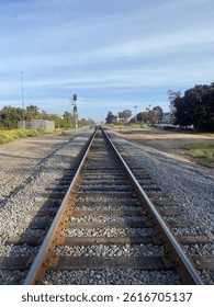 Parallel train tracks stretch into the distance under a bright, partly cloudy sky. Gravel ballast surrounds the rails, leading towards a distant signal and sparse vegetation along the sides. Linear pe