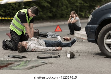 Paramedic Resuscitating On Street Car Accident Victim, In The Background Woman Talking On Phone.