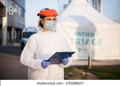 Paramedic wearing personal protective equipment PPE holding folder standing in front of ICU hospital isolation rt-PCR drive thru testing site,COVID-19 pandemic outbreak crisis,worried exhausted staff