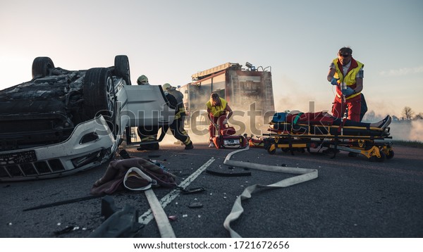 Paramedics and Firefighters Arrive On the Car Crash\
Traffic Accident Scene. Professionals Rescue Injured Victim Trapped\
in Rollover Vehicle by Extricating Them, giving First Aid and\
Extinguishing Fire