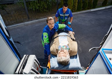 Paramedics Load Elderly Male With Breathing Mask Into Ambulance On Medical Stretcher. Emergency Treatment, EMS