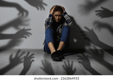 Paranoid delusion. Scared woman sitting near wall. Shadows of hands reaching for her symbolizing fear and anxiety