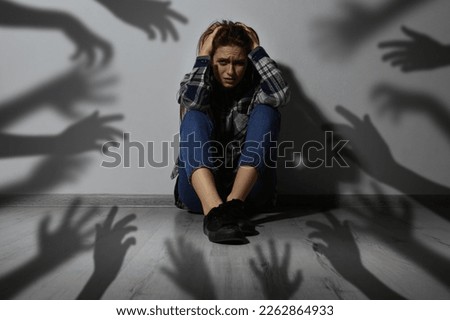 Paranoid delusion. Scared woman sitting near wall. Shadows of hands reaching for her symbolizing fear and anxiety