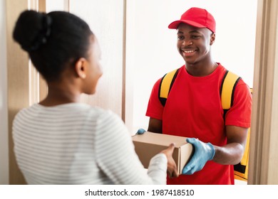 Parcel Delivery. Black Courier Man Giving Box To Lady Standing At Doors Of Her Home, Wearing Red Uniform. Woman Receiving Package Indoor. Transportation Service Concept. Selective Focus