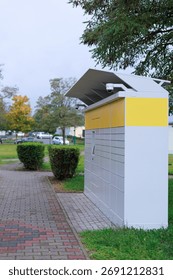 Parcel locker surrounded by grass and trees in urban park. Concept of sustainable logistics and green city solutions.