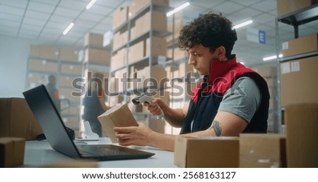 Parcel processing specialist scans package using barcode scanner, prepares for shipping to customer, works on laptop. Warehouse employees carrying boxes. E-commerce store or postal service storage.